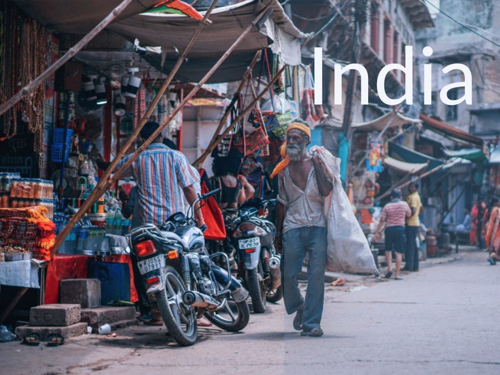 Man on street in India carrying a white bag and wearing a yellow bandana with the caption "India." Money For the Rest of Us.